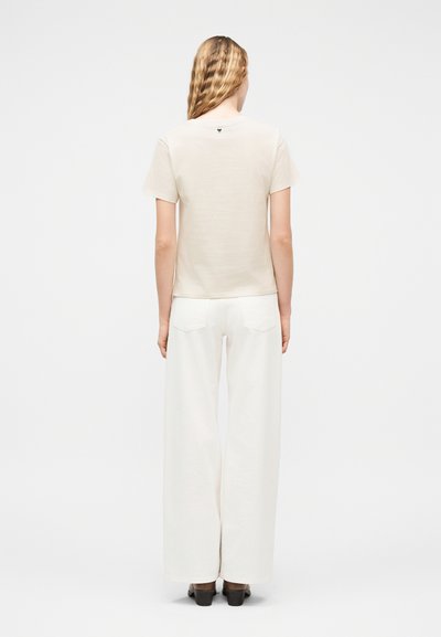 Γυναίκα με κυματιστά ξανθά μαλλιά, που φοράει μπεζ μπλούζα με κοντά μανίκια και άσπρο παντελόνι με φαρδιά πόδια, όρθια με την πλάτη προς τα εμάς σε λευκό φόντο.