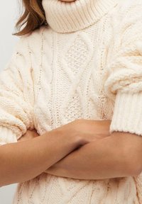 Kermanvärinen neulepaita, jossa on korkea poolokaula, teksturoidut palmikkokuviot ja hieman puffatut hihat.