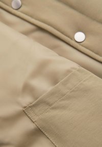 Primer plano de tela beige con costuras visibles, bordes doblados y dos botones a presión blancos en una prenda.