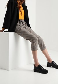 Schwarzes, übergroßes Blazerjacke, senfgelbe Bluse, karierte verkürzte Hose und schwarze Plateau-Schuhe mit Schnürsenkeln und strukturierten Details.