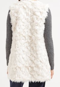 Vit fuskpälsväst med en texturerad, ruggig yta. Ärmlös design, lager ovanför en grå långärmad topp, faller nedanför höfterna.