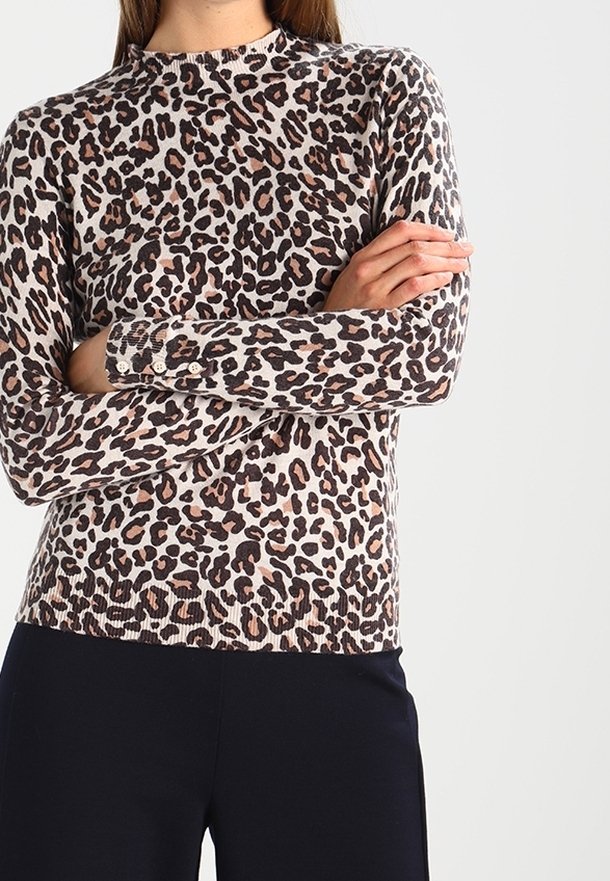 Personne portant un haut à manches longues imprimé léopard, les bras croisés, associé à un pantalon noir sur un fond uni.