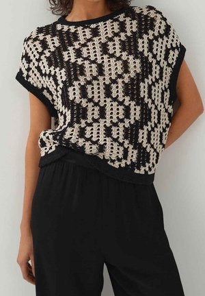 Femme portant un pull à manches courtes noir et beige à motif en maille ajourée et un pantalon noir ample, debout devant un fond uni.