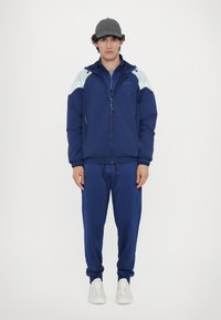 Marineblå vindjakke med lyseblå detaljer, lynlås lukning og to sidelommer; parret med matchende marineblå sweatsbukser og hvide sneakers.