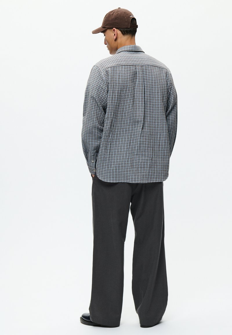 Man wearing brown cap, blue and white checkered shirt, dark loose pants, and black shoes, standing facing away on a white background.