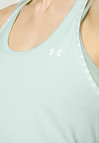 Vihreä urheilullinen hihaton paita pyöreällä kaula-aukkolla ja valkoisella Under Armour -logolla. Mukana on hillittyä tekstiä hihassa. Sileä pinta.