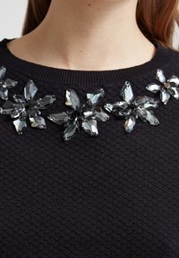 Czarny sweter z teksturowanej tkaniny z kwiecistym wzorem z ciemnych kamieni szlachetnych wokół dekoltu.