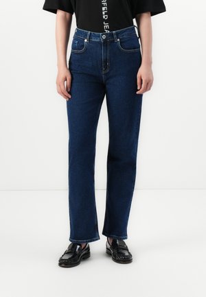 Dark blue denim jeans with a straight leg design, high waist, five pockets, and silver hardware; worn with a black t-shirt and loafers.