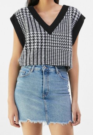 Vrouw draagt een zwart-wit pied-de-poule gebreid mouwloos vest met diepe V-hals en een lichtblauwe denim minirok met rafels.