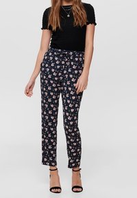 Pantalon bleu à motifs floraux avec des fleurs roses, tissu léger, coupe droite, avec une taille haute et un détail noué. Associé à un haut noir.