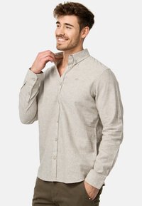 Uomo sorridente che aggiusta il colletto di una camicia beige con bottoni e maniche lunghe, infilata in pantaloni verde oliva, in piedi davanti a uno sfondo semplice.