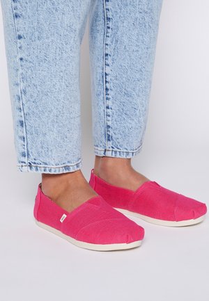 Voeten met felroze instapcanvas schoenen gecombineerd met lichtblauwe cropped spijkerbroeken tegen een effen achtergrond.