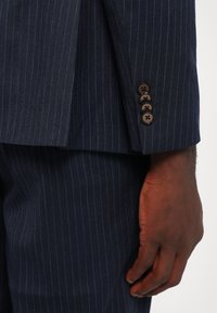 Ingrandimento di una mano di carnagione scura accanto alla manica di un giubbotto blu navy a righe con quattro bottoni marroni.