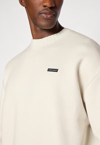 Sweatshirt de couleur crème avec un col rond, des poignets côtelés et une petite étiquette noire affichant le nom de la marque à l'avant.