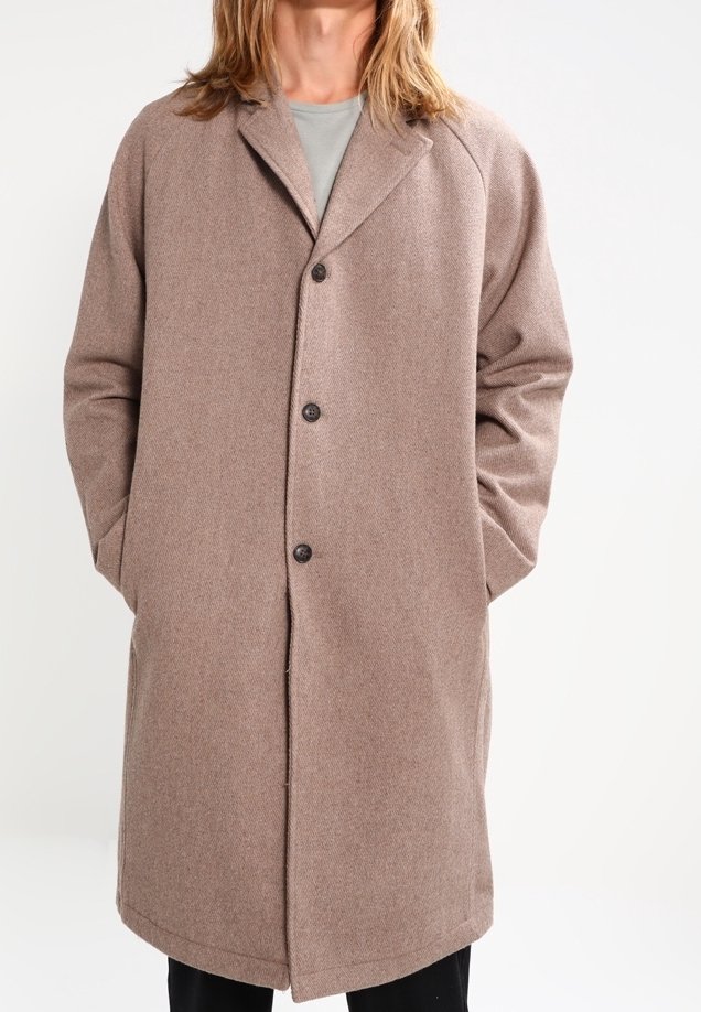 Personne portant un manteau en laine taupe boutonné jusqu'aux genoux avec des poches latérales et une chemise gris clair en dessous.