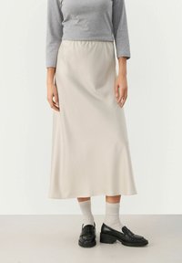 Keskipituinen, beige satiinihame, joka on sileäpintainen, yhdistettynä harmaaseen yläosaan ja mustiin loafer-kenkiin, joita täydentävät vaaleanharmaat sukat.