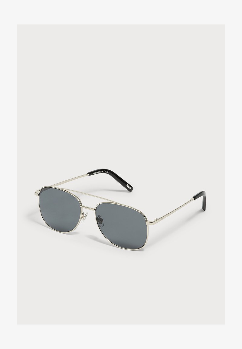 Lunettes de soleil aviateur en métal argenté avec verres teintés foncés et embouts de branches noirs sur fond clair.