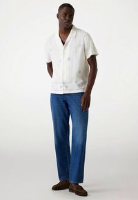 Košulja svijetle boje s kratkim rukavima i plavim cvjetnim uzorkom, u kombinaciji s plavim trapericama i smeđim cipelama u stilu mokasinki.