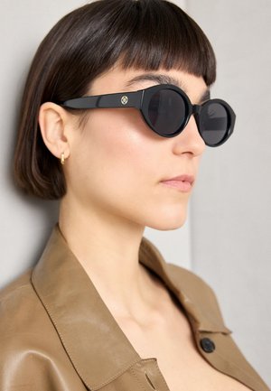 Mustat oktaaliset aurinkolasit tummilla linsseillä, joissa on kultainen emblem sivulla, yhdistettynä vaaleanruskeaan nahkatakkiin.