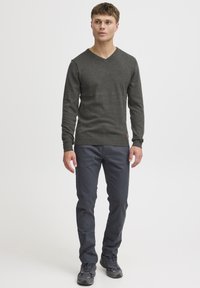 Pull en V noir en charbon avec des poignets et un ourlet côtelés, associé à un pantalon slim-fit bleu marine et des chaussures de sport noires, se tenant devant un fond blanc.