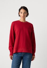 Roter Baumwollpullover mit rundem Ausschnitt, langen Ärmeln und gerippten Bündchen. Mit einer gestickten Detail auf der Brust und einem Etikett am Ärmel.