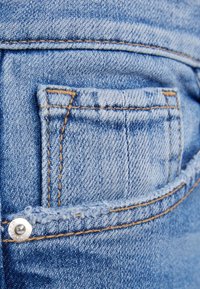 Tejido de denim azul claro con un pequeño bolsillo trasero. Presenta costuras doradas y un detalle de botón metálico. La textura parece ligeramente desgastada.