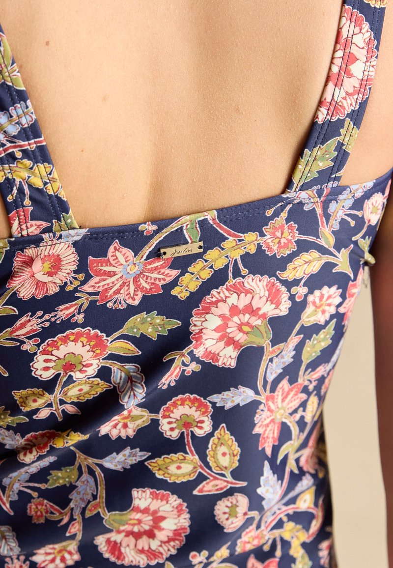 Nahaufnahme des oberen Rückens einer Person, die ein dunkelblaues ärmelloses Top mit einem bunten Blumenmuster und einem kleinen Metallmarkenetikett trägt.