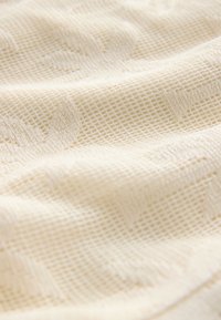 Tessuto di colore crema con texture e un motivo tessuto, caratterizzato da un design a griglia e sottili forme di foglie, che mette in risalto morbidezza e profondità.