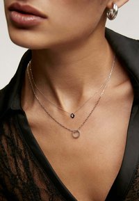 Collares de plata apilados que presentan un colgante circular con pequeños acentos, un charm en forma de corazón y una cadena fina. Tela negra texturizada en el fondo.