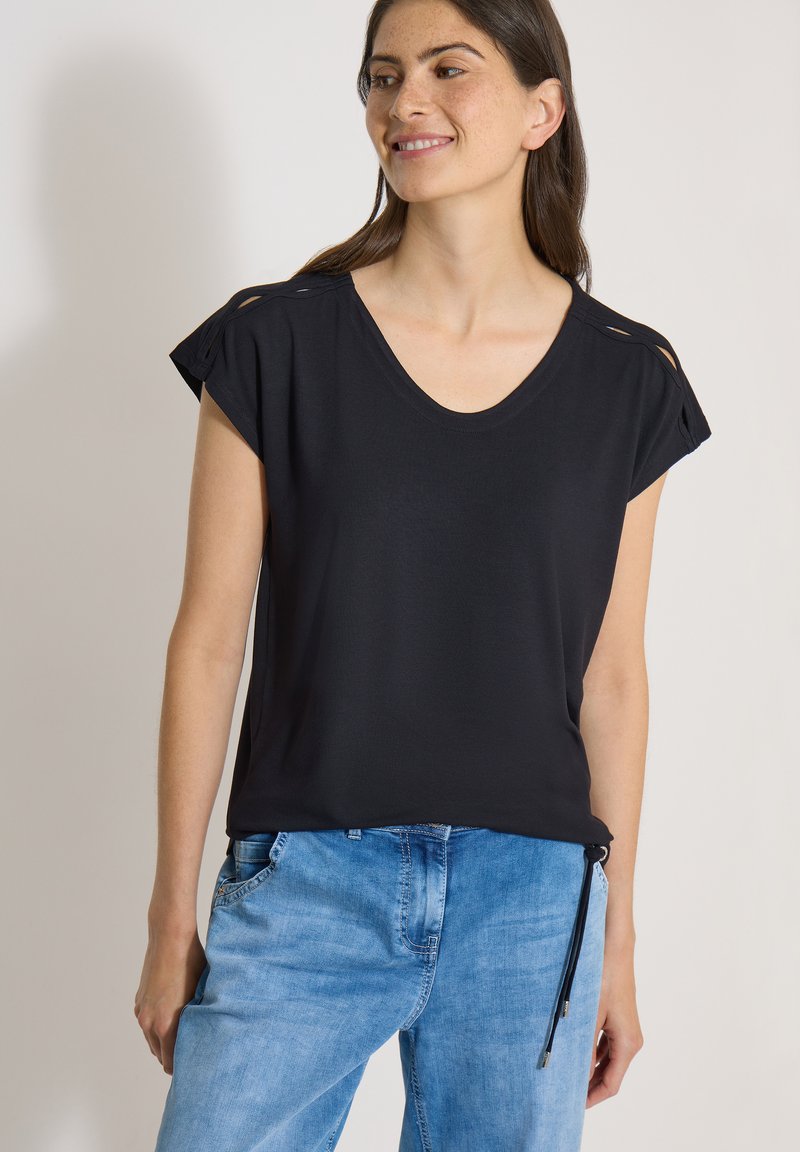 Femme portant un haut noir à manches courtes avec des découpes aux épaules et un jean en denim bleu clair sur un fond uni.