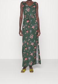 Blommig maxi-klänning i grönt tyg med knytning i midjan, prydd med rosa och blå blommor, V-ringning och en sidoöppning. Kombinerad med gröna klackar.