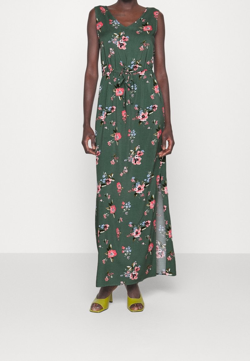 Blommig maxi-klänning i grönt tyg med knytning i midjan, prydd med rosa och blå blommor, V-ringning och en sidoöppning. Kombinerad med gröna klackar.