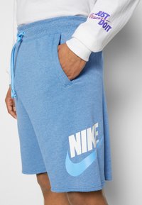 Blaue Fleece-Shorts mit elastischem Bund und Kordelzug. Großes weißes "NIKE"-Logo mit einem hellblauen Swoosh auf der linken Seite.