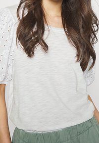 Bílý bavlněný top s texturovanou tkaninou a nadýchanými bílými krajkovými rukávy s květinovými výřezy, v kombinaci se zelenými kalhotami s elastickým pasem.