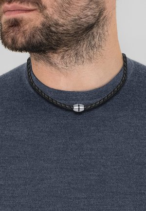 Collier en cuir tressé noir avec un fermoir en métal argenté, porté autour du cou sur un pull bleu foncé.