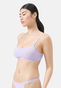 Frau mit langen, dunklen Haaren, die nach hinten gebunden sind, trägt einen helllila Bralette und passende Unterwäsche, blickt nach links vor einem schlichten Hintergrund.