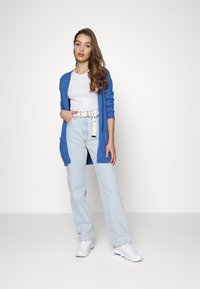 Cardigã tricotado azul royal, t-shirt branca justa, jeans de perna larga azul clara, ténis brancos. O cardigã tem frente aberta e bolsos.