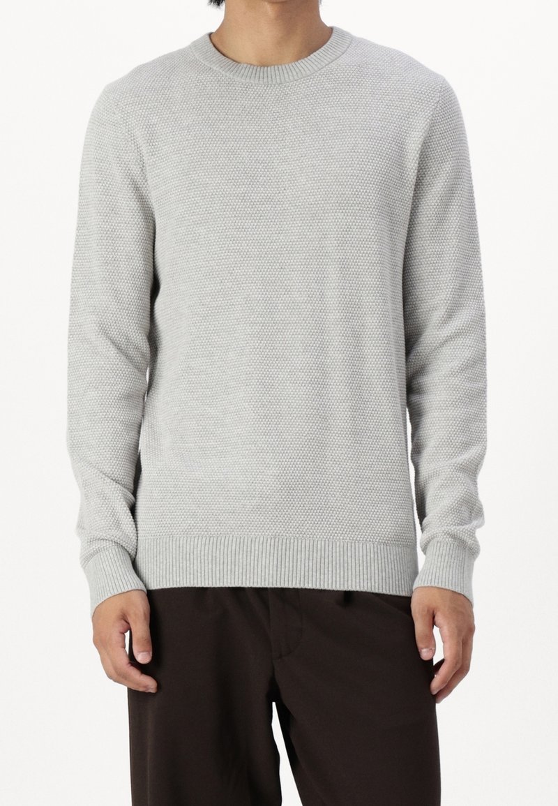 Maglione grigio chiaro lavorato a maglia con un motivo strutturato, colletto rotondo e polsini a coste. Indossato con pantaloni marroni scuri.