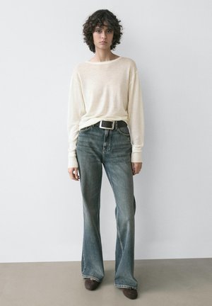 Junge Person mit lockigem Haar, die ein cremefarbenes langärmliges Oberteil, hoch taillierte, verwaschen blaue Jeans, schwarzen Gürtel und dunkle Schuhe trägt und an einer Wand steht.