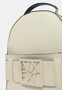 Mochila color crema hecha de material sintético texturizado, con un parche de logo rectangular y detalles en negro en los bordes.