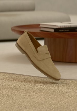 Beige suede loafer shoe floating above beige woven rug, with round wooden coffee table and stacked books in the background.