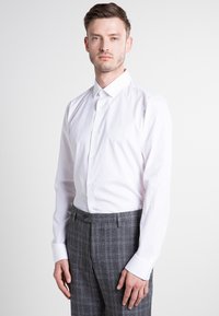 Mann in einem weißen Hemd mit Knöpfen und grauen karierten Hosen, der vor einem schlichten weißen Hintergrund steht und nach vorne blickt.