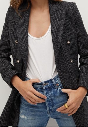 Frau trägt einen dunkelgrauen karierten Blazer über einem weißen T-Shirt mit Rundhalsausschnitt, das in hoch taillierte blaue Jeans mit Used-Details gesteckt ist.