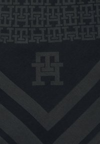 Mørkt stof med sammenflettede geometriske "H"-mønstre og stort centreret "HH"-logo over chevron-striber i kontrasterende nuancer.