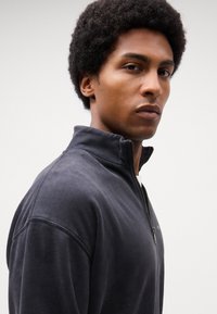 Mørkegrå zip-up sweatshirt med høj krave, afslappet pasform og struktureret stof, der har en synlig lynlås og diskrete syninger.