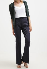 Marina skräddarsydda byxor med utställd design, kombinerade med en vit t-shirt med v-ringning och en mörkgrön kofta. Sandaler fulländar outfiten.