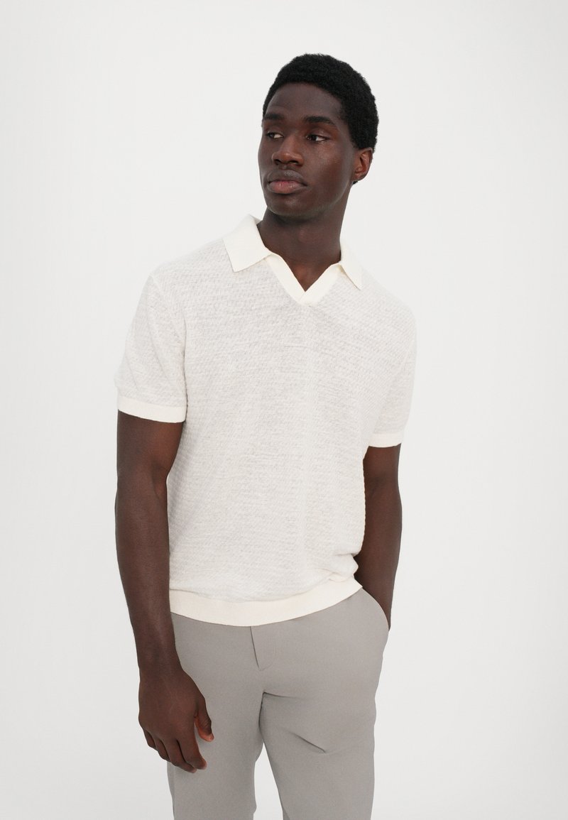 Short-sleeved, v-neck polo shirt in light cream, textured material with a contrasting white collar and ribbed hem, paired with light gray trousers.