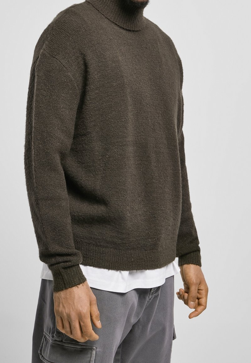 Homme portant un pull à col roulé marron foncé sur une chemise blanche et un pantalon cargo gris sur un fond clair uni.