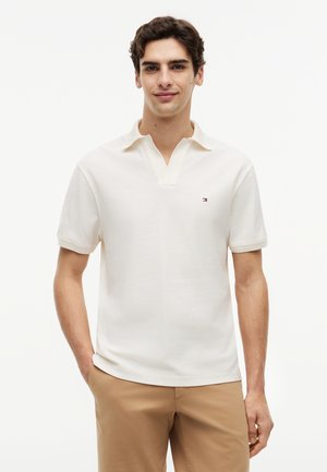 Νεαρός άνδρας με κοντά σκουρόχρωμα μαλλιά, ντυμένος με λευκό πόλο με κοντά μανίκια και μπεζ παντελόνι, στέκεται με το ένα χέρι στην τσέπη.