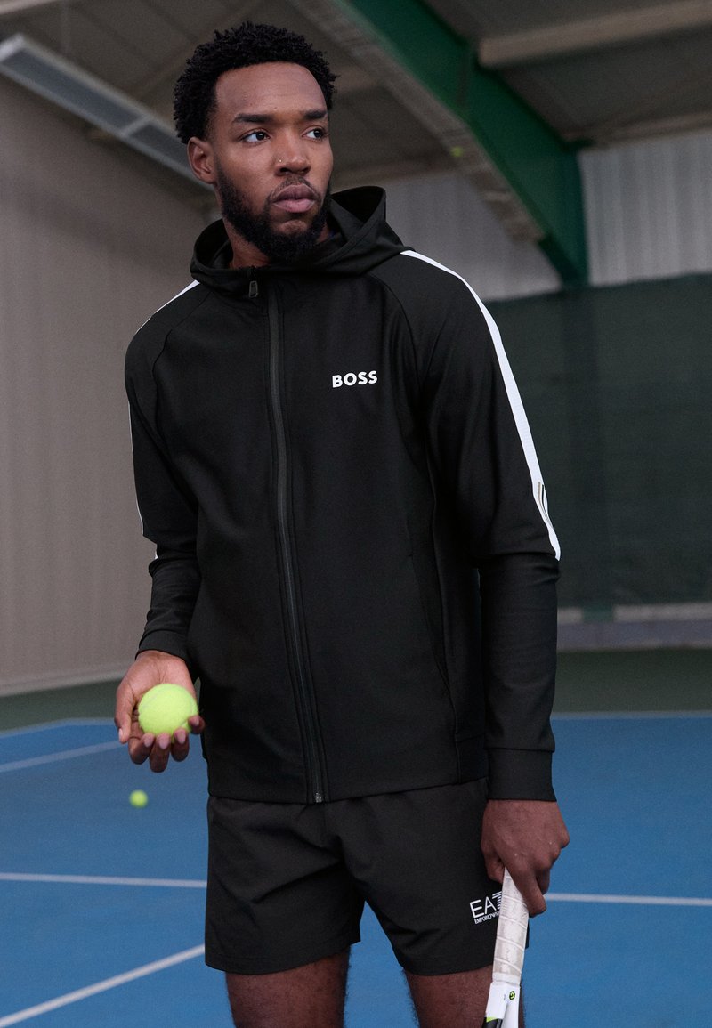 Schwarzer sportlicher Kapuzenpullover mit Reißverschluss, weißen Akzenten und dem "BOSS"-Logo. Kombiniert mit schwarzen Shorts, hält einen Tennisball und einen Schläger.